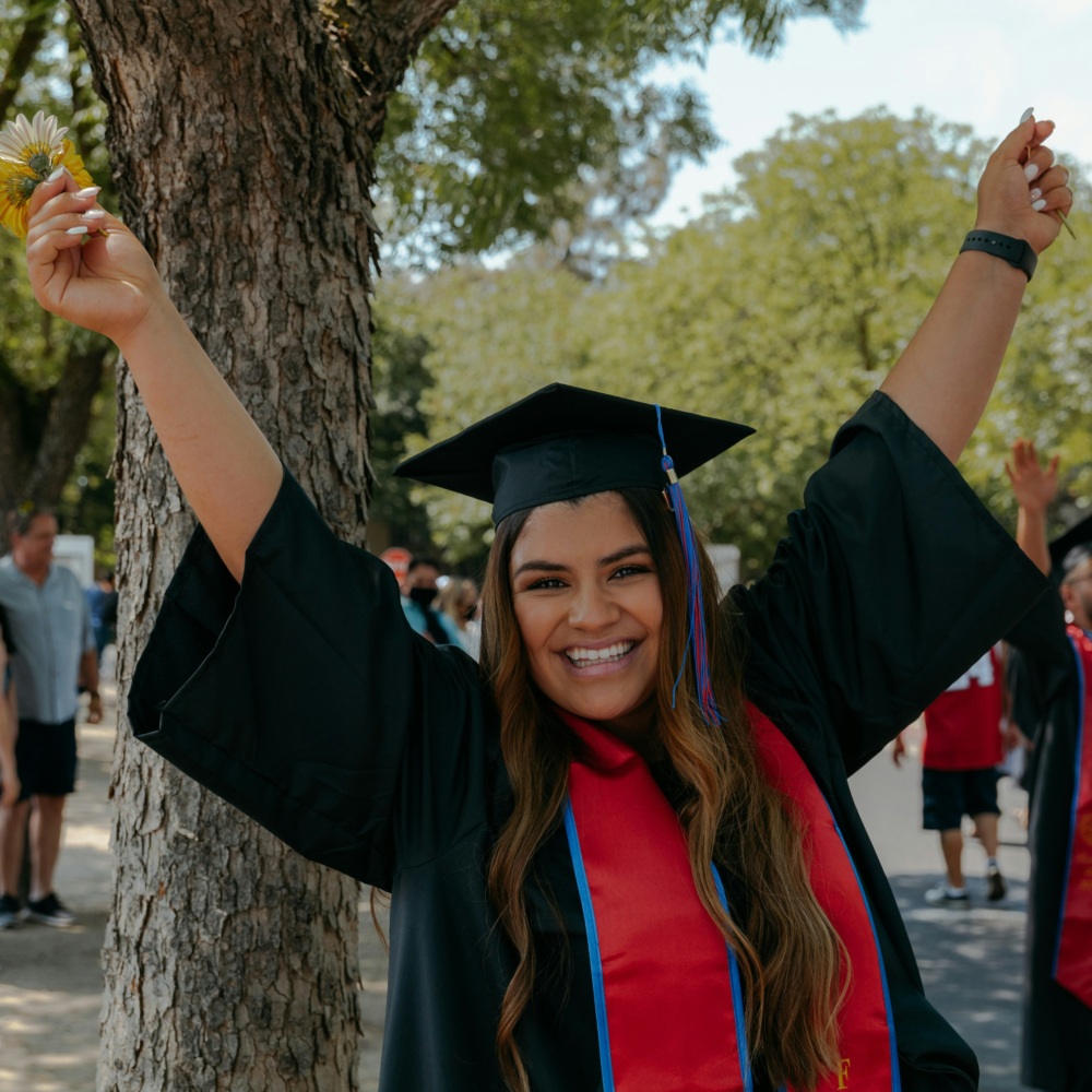 A smiling woman wearing a graduation cap and gown raises her arms in celebration beside a tree.